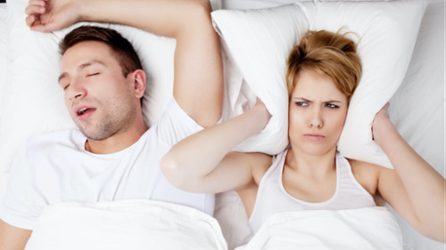 Man snoring in bed while frustrated partner covers ears with pillow